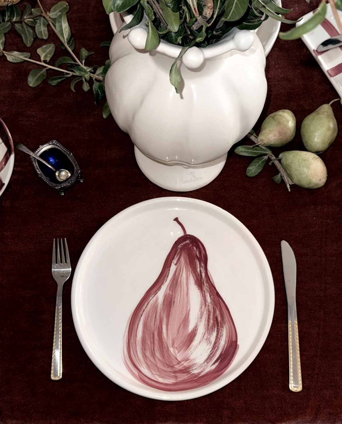 A white ceramic dinner plate featuring a painted red pear design, styled on a dark red tablecloth with a silver fork, knife, white vase with greenery, and pears.