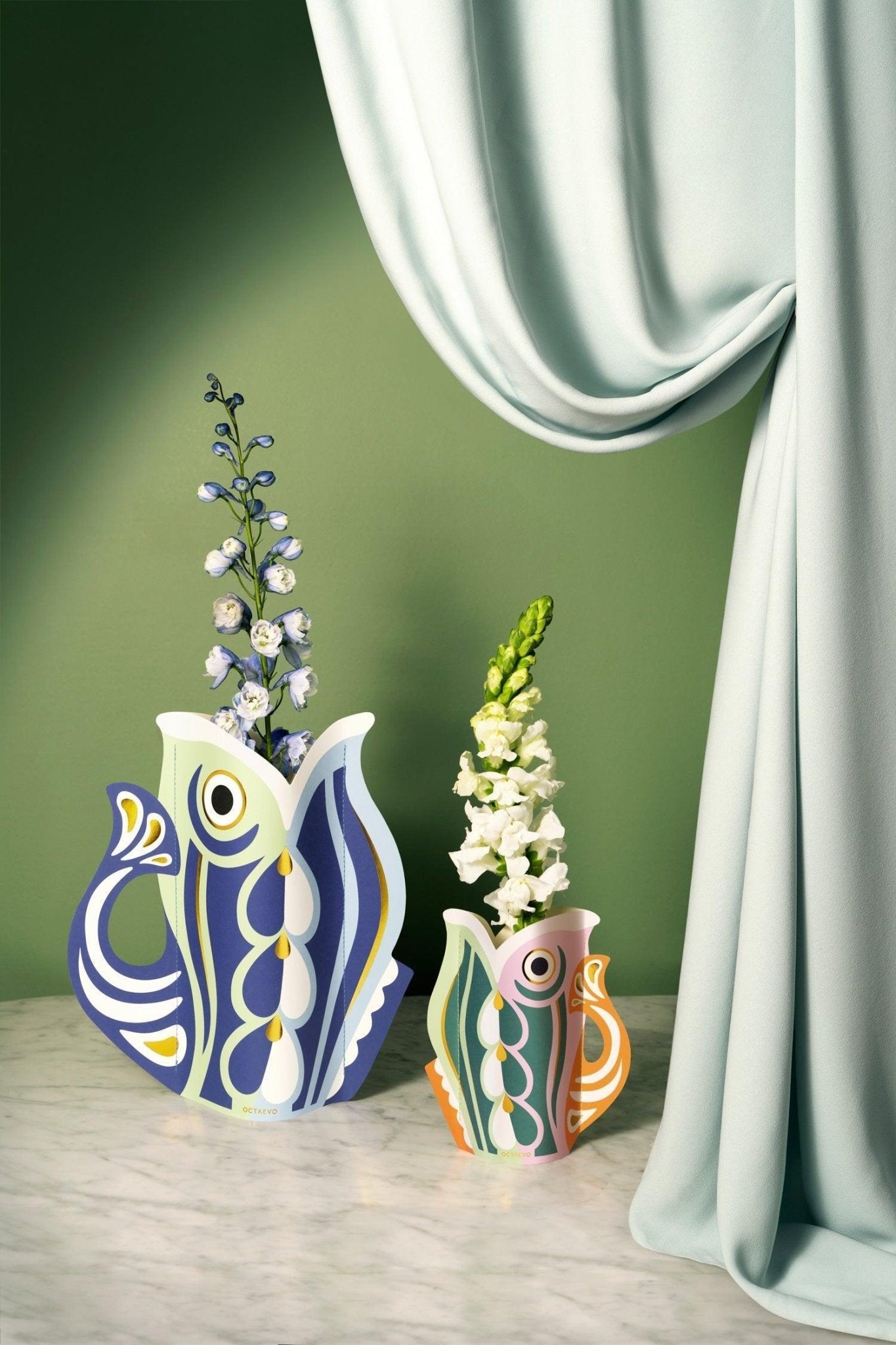 Two decorative OCTAEVO paper vases in blue with floral arrangements against a green curtain background.
