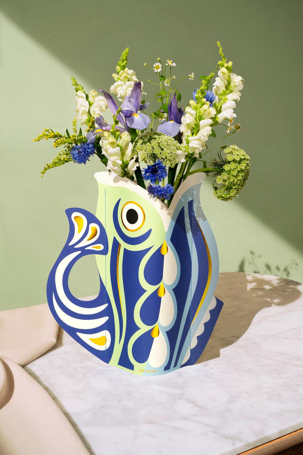Decorative blue OCTAEAVO fish-shaped paper vase with flowers against a green wall. 