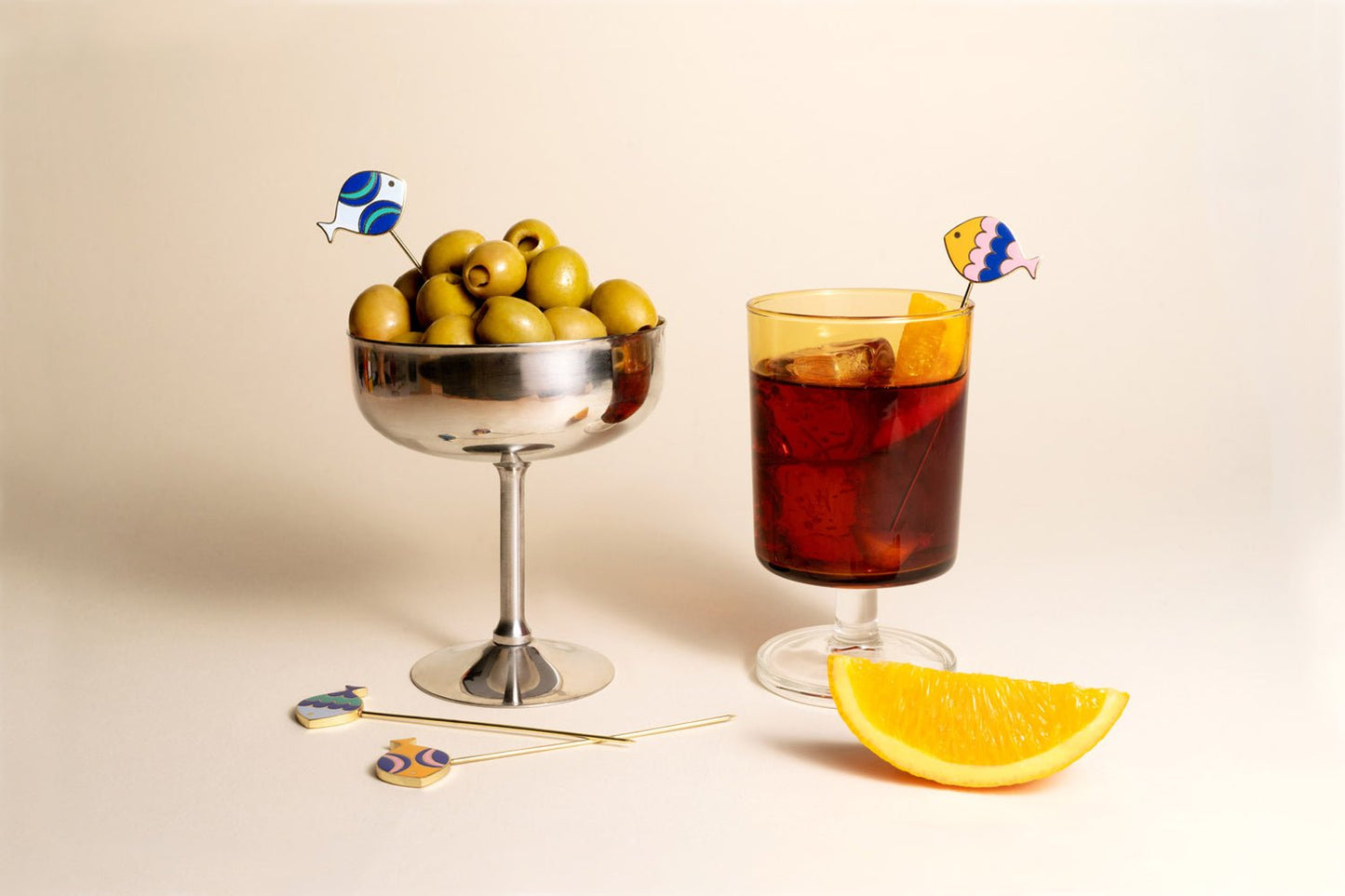 A glass cup of olives and a red iced drink with orange, adorned with Octaevo Fish Splash Cocktail Picks.