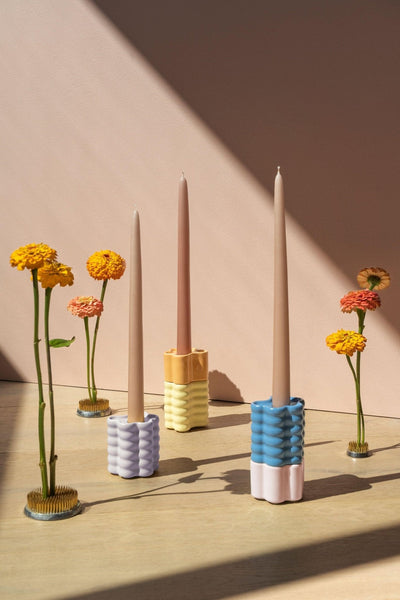 Three Octaevo Flora Candle Holders in pink with tall candles are arranged among flowers.