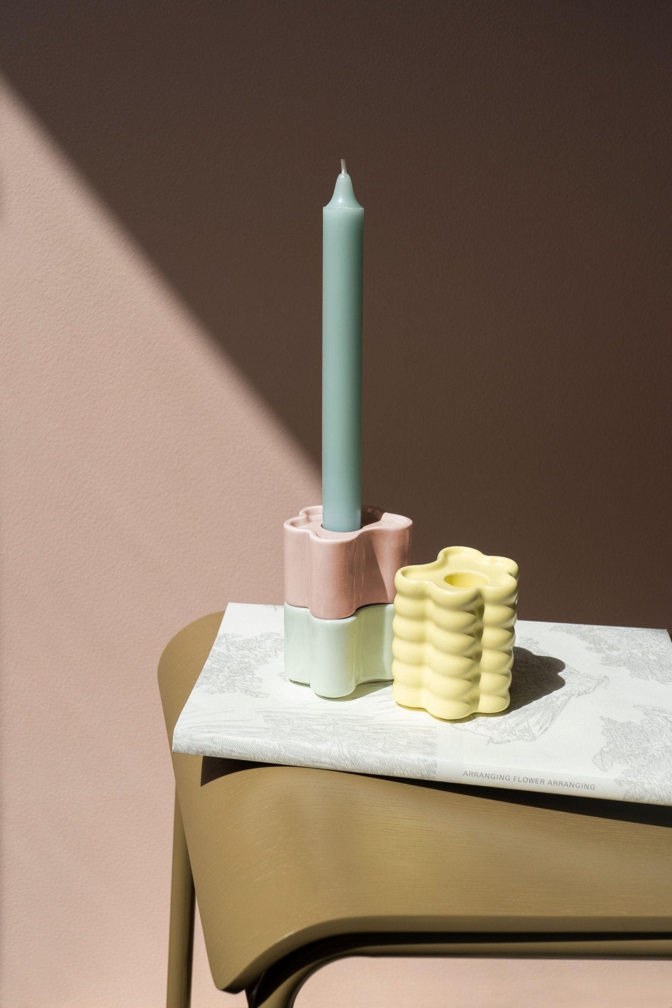A pastel blue candle in an Octaevo Flora Candle Holder in mint rest on a book atop a tan table under soft light.
