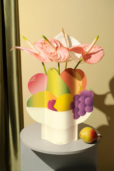 The Octaevo Fruit Bowl Paper Vase with pink anthuriums and fruit shapes is a vibrant accent on a round gray pedestal.