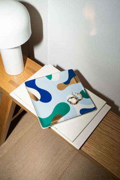 The Gaia Ceramic Tray by Octaevo holds gold hoop earrings and shines with gold foil, a chic accent beside books and a lamp.