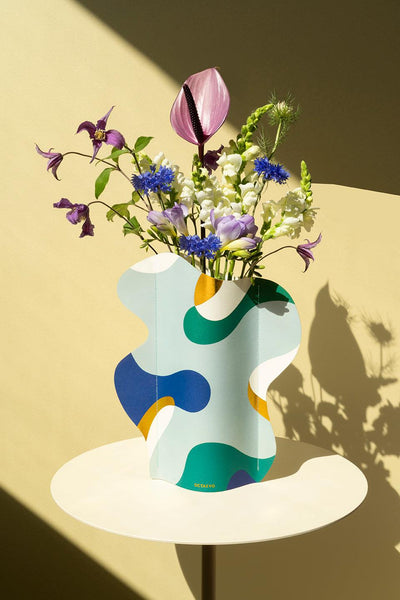 The Gaia Paper Vase by Octaevo with fresh flowers sits on a round white table, casting a shadow on a beige wall.