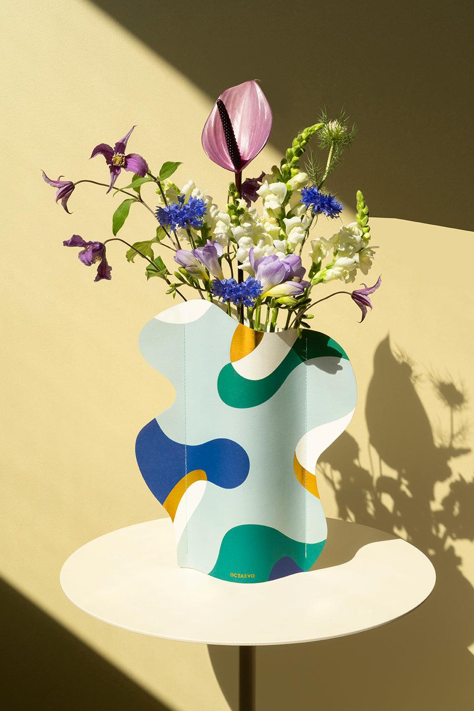 The Gaia Paper Vase by Octaevo with fresh flowers sits on a round white table, casting a shadow on a beige wall.