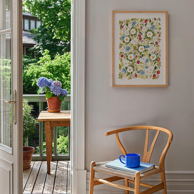Garden Strawberries Print in light wood frame, styled A2 Print against white wall in kitchen with chair, and flower pots outside.