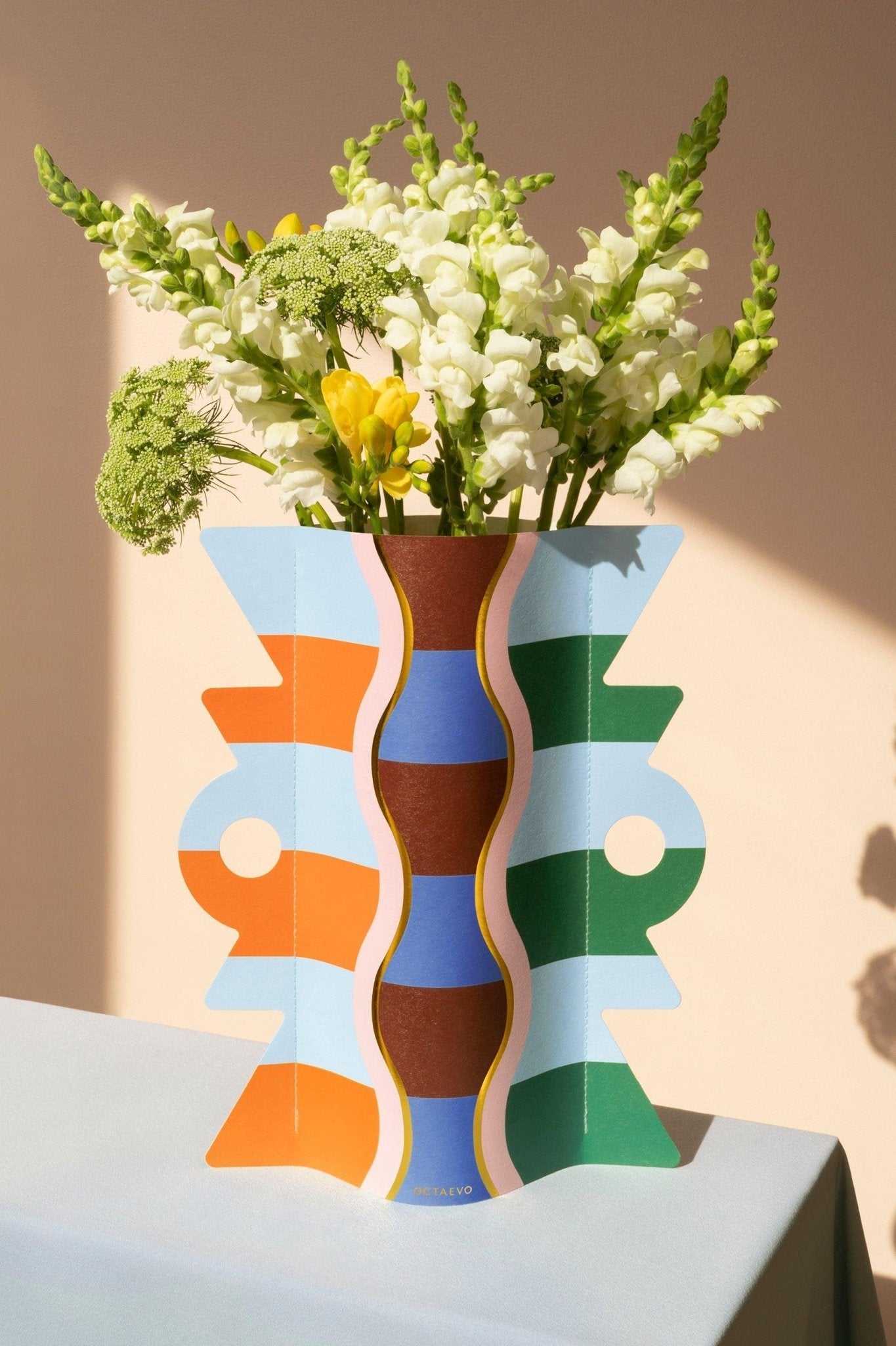 The Giza Paper Vase by OCTAEVO holds white and yellow flowers on a light blue table with a beige background.