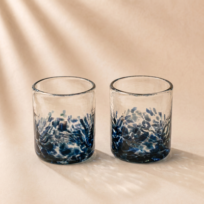 Two clear glass tumblers with a textured, hammered blue pattern concentrated at the bottom, set against a warm beige background with soft leaf shadows.