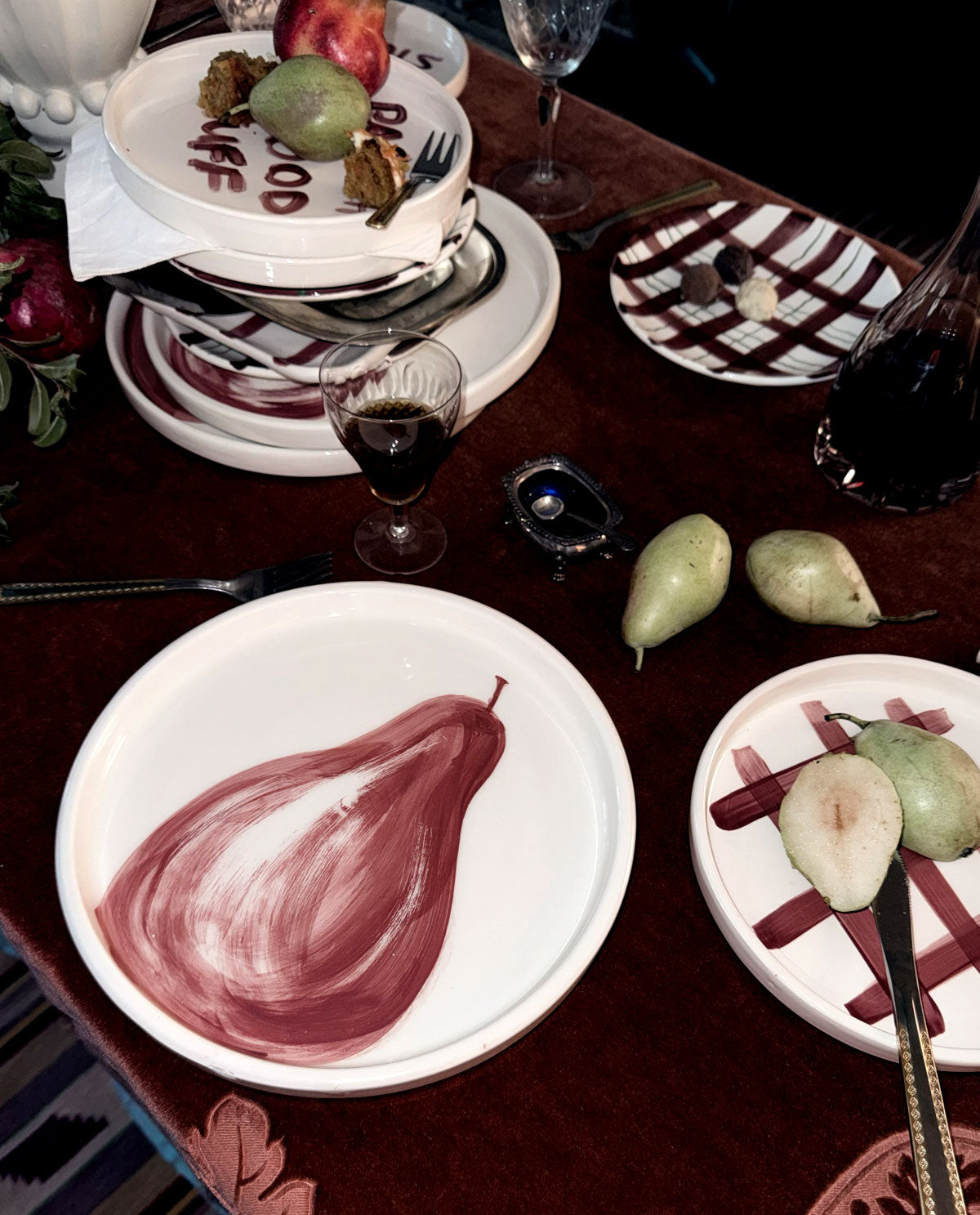 A tabletop dining scene with white plates featuring painted pear motifs, wine glasses, fruit, and a dark red tablecloth in a styled indoor setting.