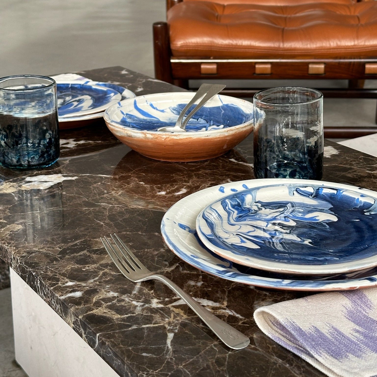 Dining table setting with blue and white dishes, glasses, and cutlery on a marble surface with a brown leather chair in the background - lifestyle
