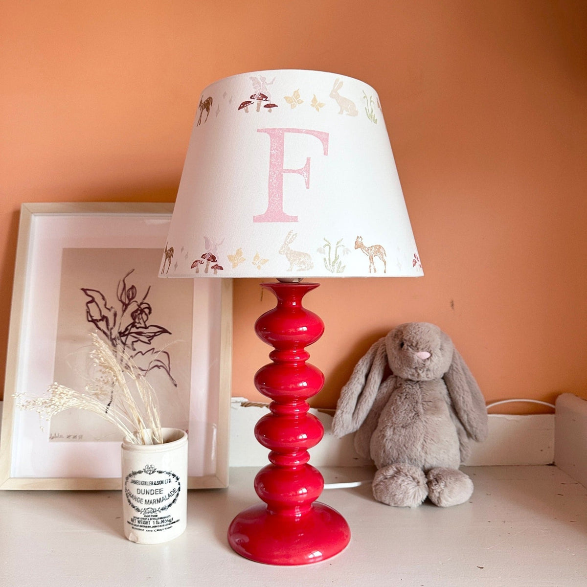 Personalised kids lampshade with woodland animals design and pink initial, styled on a red lamp base in a girl’s bedroom. A pretty empire lampshade ideal for kids bedroom décor and new parent gifts.
