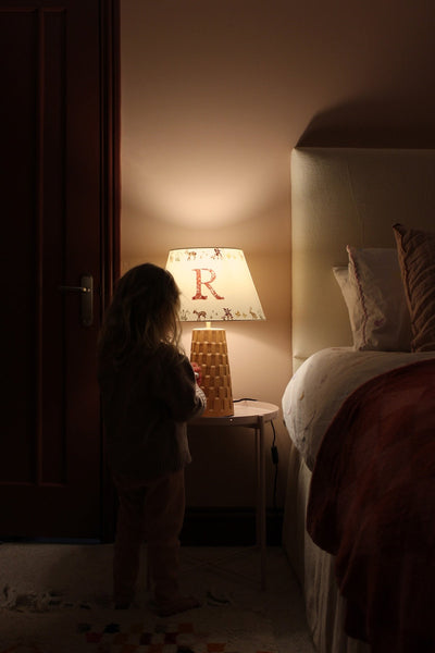 A dimly lit bedroom with a glowing personalised lampshade featuring and a woodland pattern. A young girl stands beside it, reaching for the lamp.
