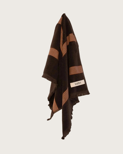 Chocolate brown hand towel with fringing and red and cream stripes hanging on a neutral background. 