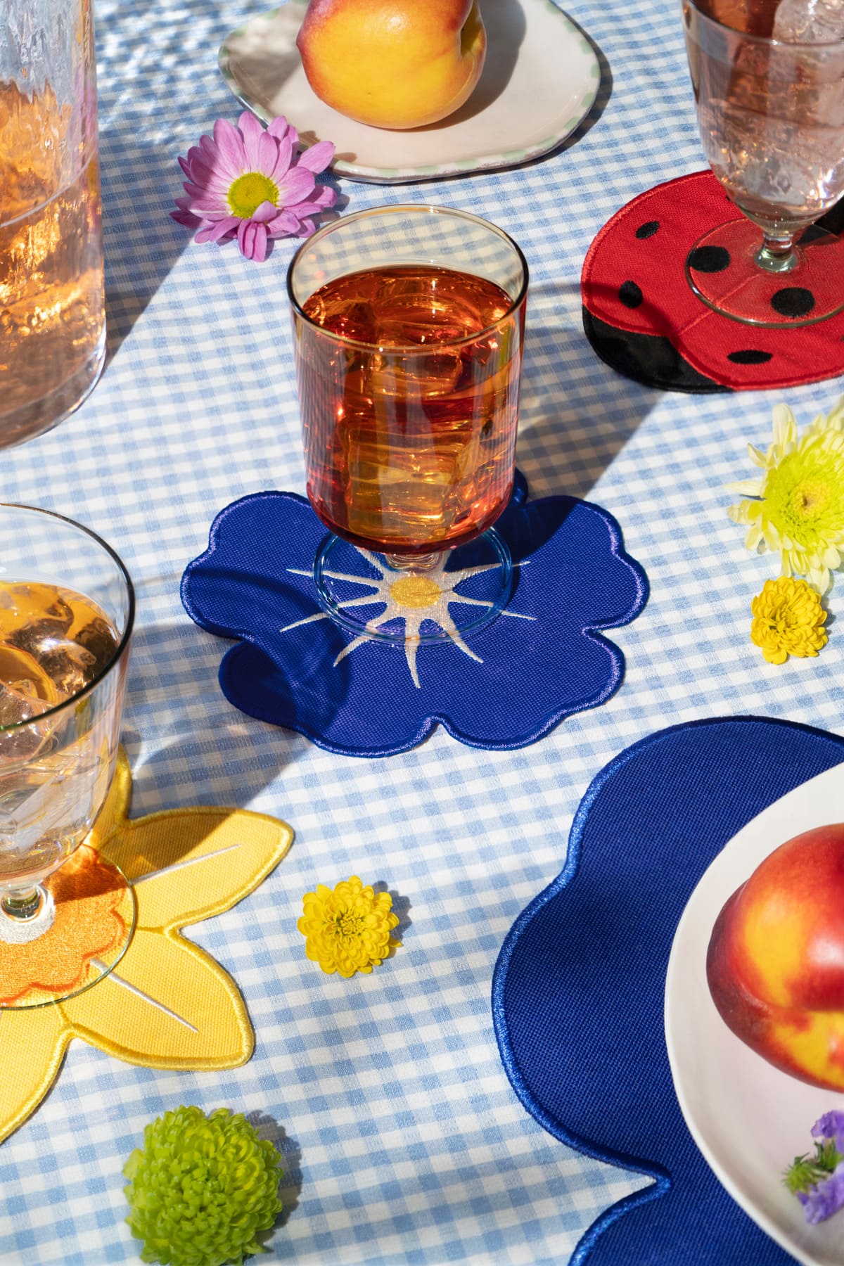 A glass of amber liquid on Octaevo Ladybug Coasters atop a blue gingham tablecloth with flowers, plates, and drinks.