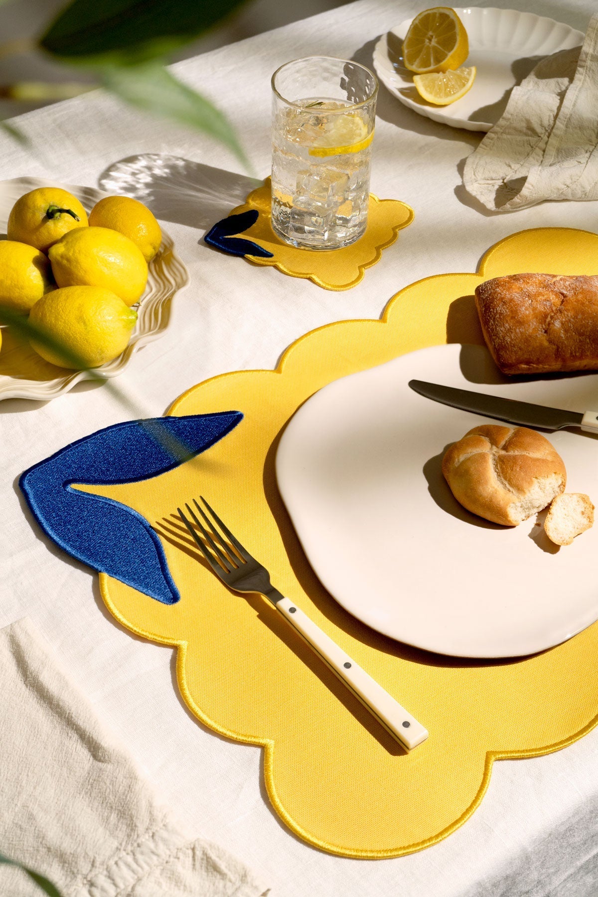 A sunny table with lemons, bread, yellow placemats, and Octaevo Lemon Coasters (Set of 4) on a white linen-cotton cloth.