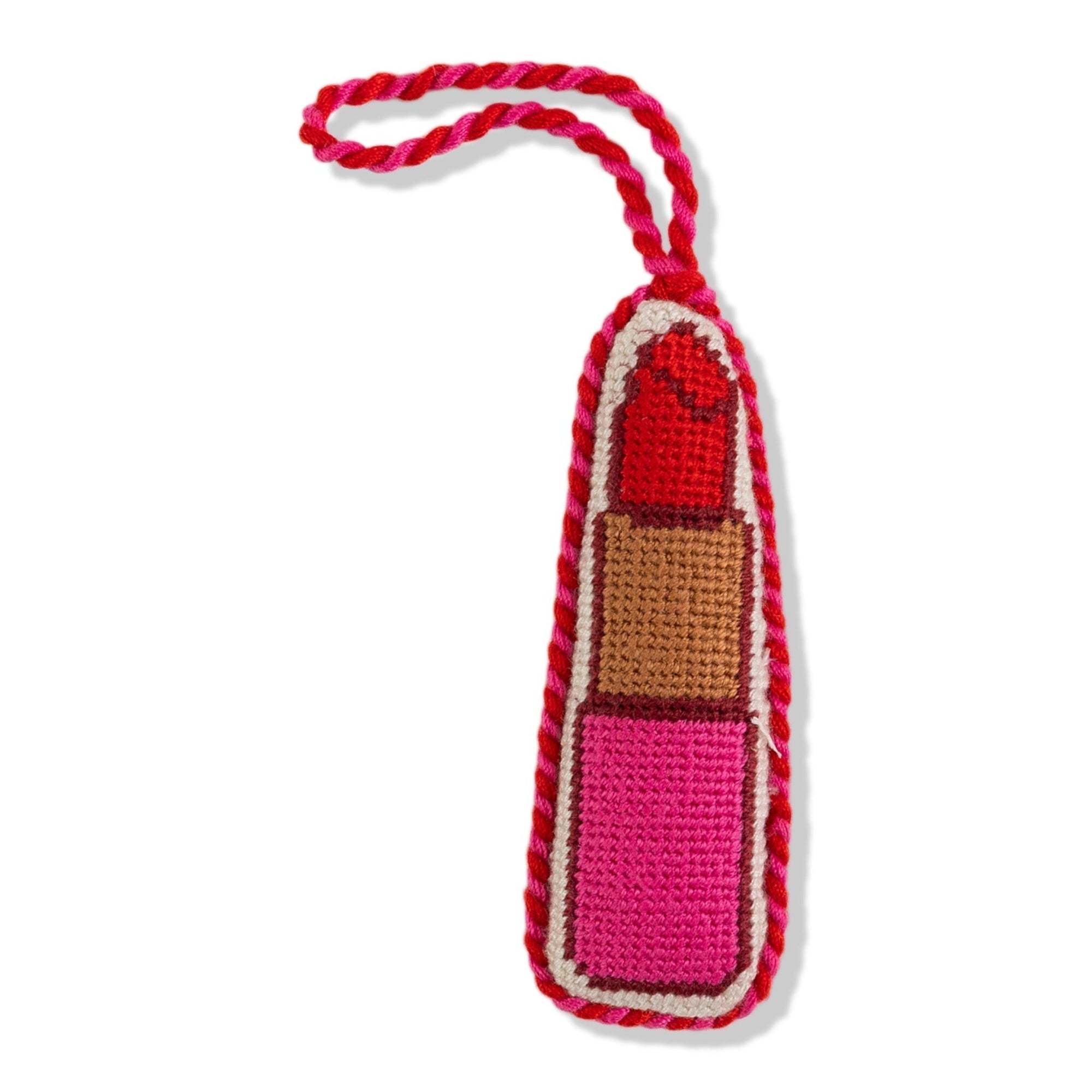 A needlepoint ornament shaped like a lipstick with a red, beige, and pink design, backed with red velvet and tied with a red ribbon.