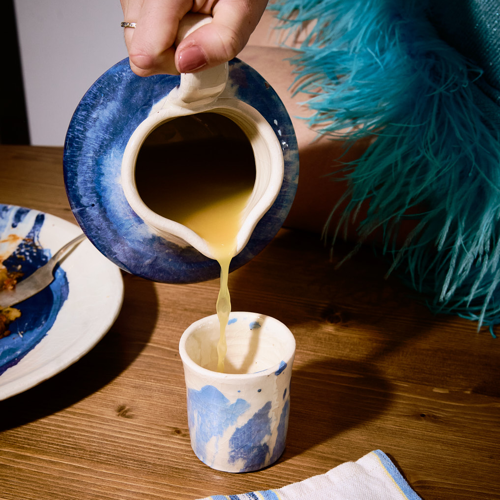 Person pouring a liquid from a blue and white ceramic pitcher into a matching cup on a wooden table.
