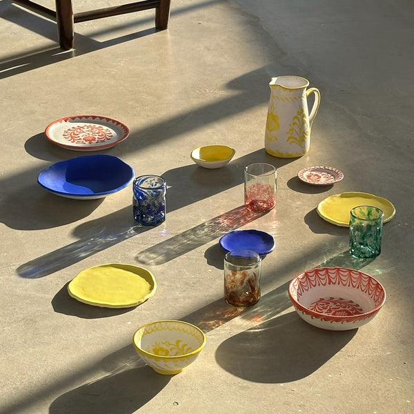 Colorful ceramic dishes and glasses arranged on a concrete floor with a brown leather ottoman in the background. - lifestyle