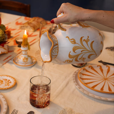 Person pouring liquid from a decorative teapot into a glass on a table set with plates and candles. - lifestyle