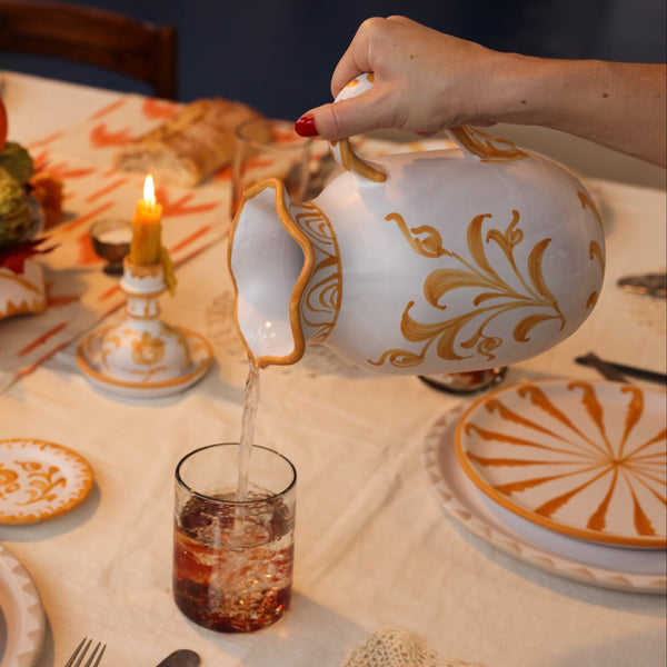 Person pouring liquid from a decorative teapot into a glass on a table set with plates and candles. - lifestyle