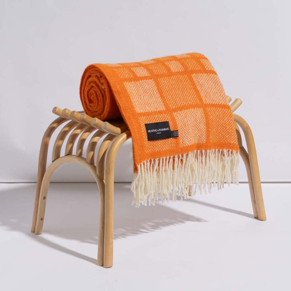 Bold orange wool blanket with white square patterns and fringed edges, neatly rolled on a wooden bench—luxury throw for indoor decor.