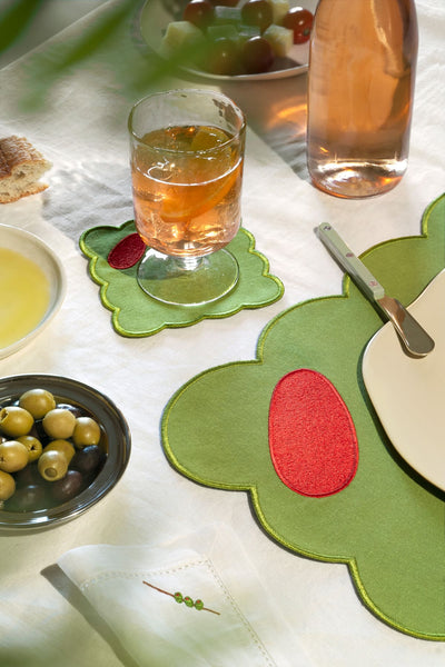 A glass of iced drink on Octaevo’s Olive Coasters (Set of 4), with plates of olives and grapes on playful placemats.