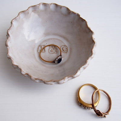 Ceramic dish with 'M & G' branding holding a gold ring, on a white background.