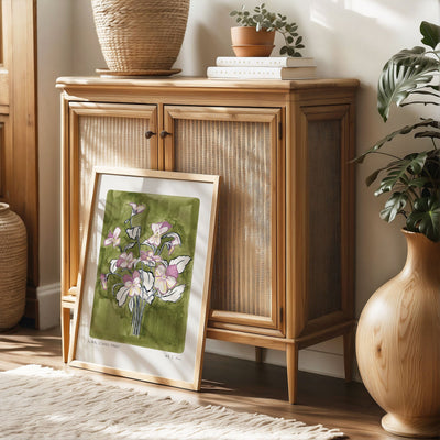 Pansy Art Print, a olive green painted background with light purple and light yellow pansy flower heads with navy outlined leaves. With small text reading Floral studies: pansy NO.1 styled in a light wood frame against rattan furniture