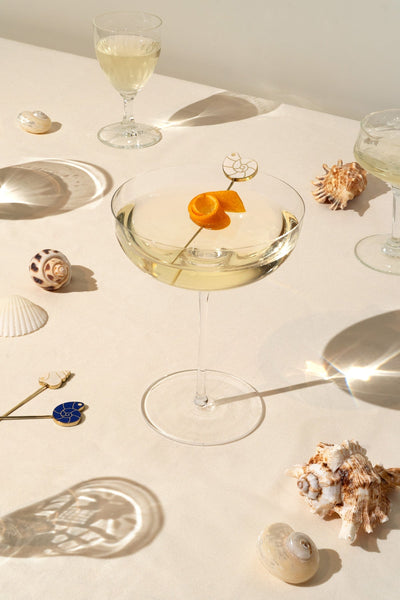 A cocktail glass with a citrus twist and OCTAEVO Pearl Shell Cocktail Picks (Set of 4) on a sunlit, seashell-adorned table.