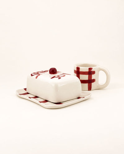 A ceramic butter dish with a lidded container and a matching mug, both in white with dark red plaid and script details, photographed on a clean off-white background.