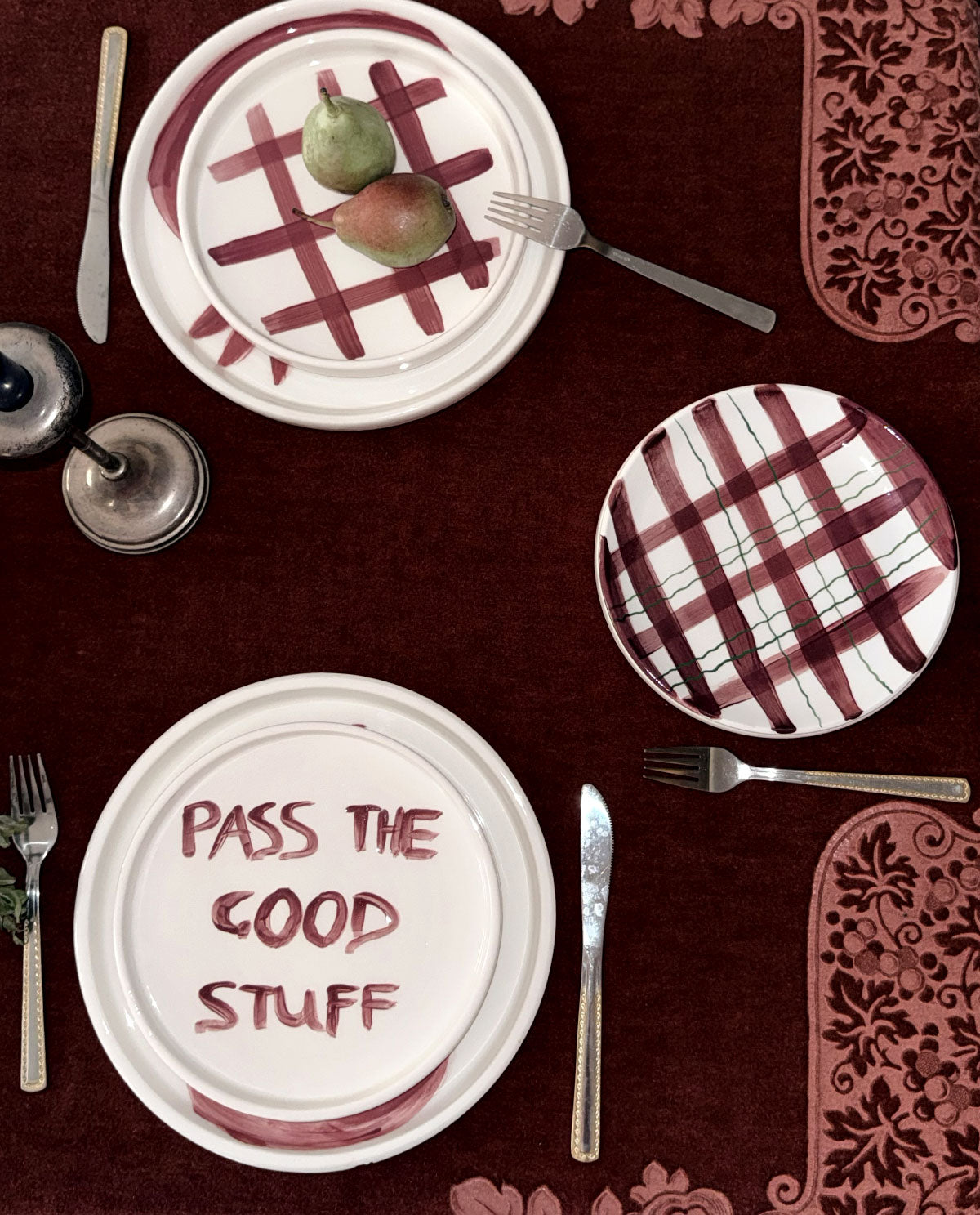 Three white ceramic plates with burgundy patterns, set on a dark red tablecloth with silver cutlery. One plate features the phrase 'PASS THE GOOD STUFF' in burgundy lettering, and two other plates display burgundy plaid and checkered designs.