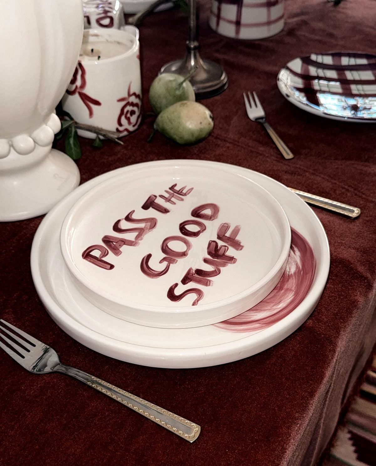 A white ceramic dinner plate set on a dark red tablecloth, featuring a stack of plates with the phrase 'PASS THE GOOD STUFF' written in red across the top plate. Silver-toned forks and other tableware are arranged around it.