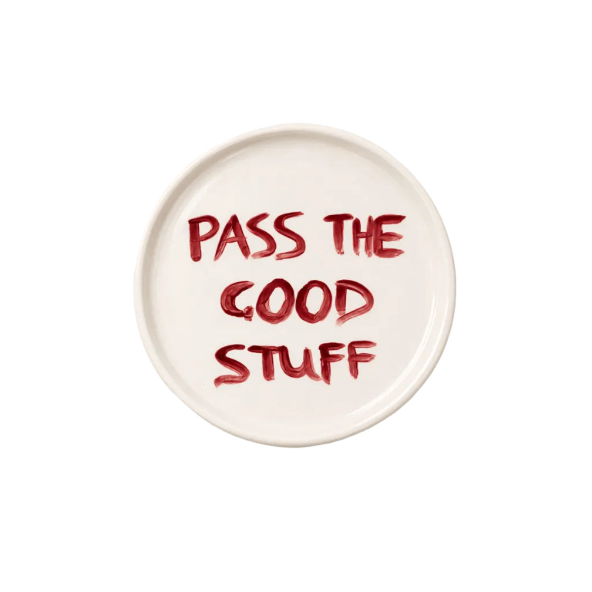 A round white ceramic plate with the phrase 'PASS THE GOOD STUFF' written in a textured red brush-style font, photographed from directly above on a clean white background.