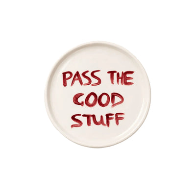 A round white ceramic plate with the phrase 'PASS THE GOOD STUFF' written in a textured red brush-style font, photographed from directly above on a clean white background.