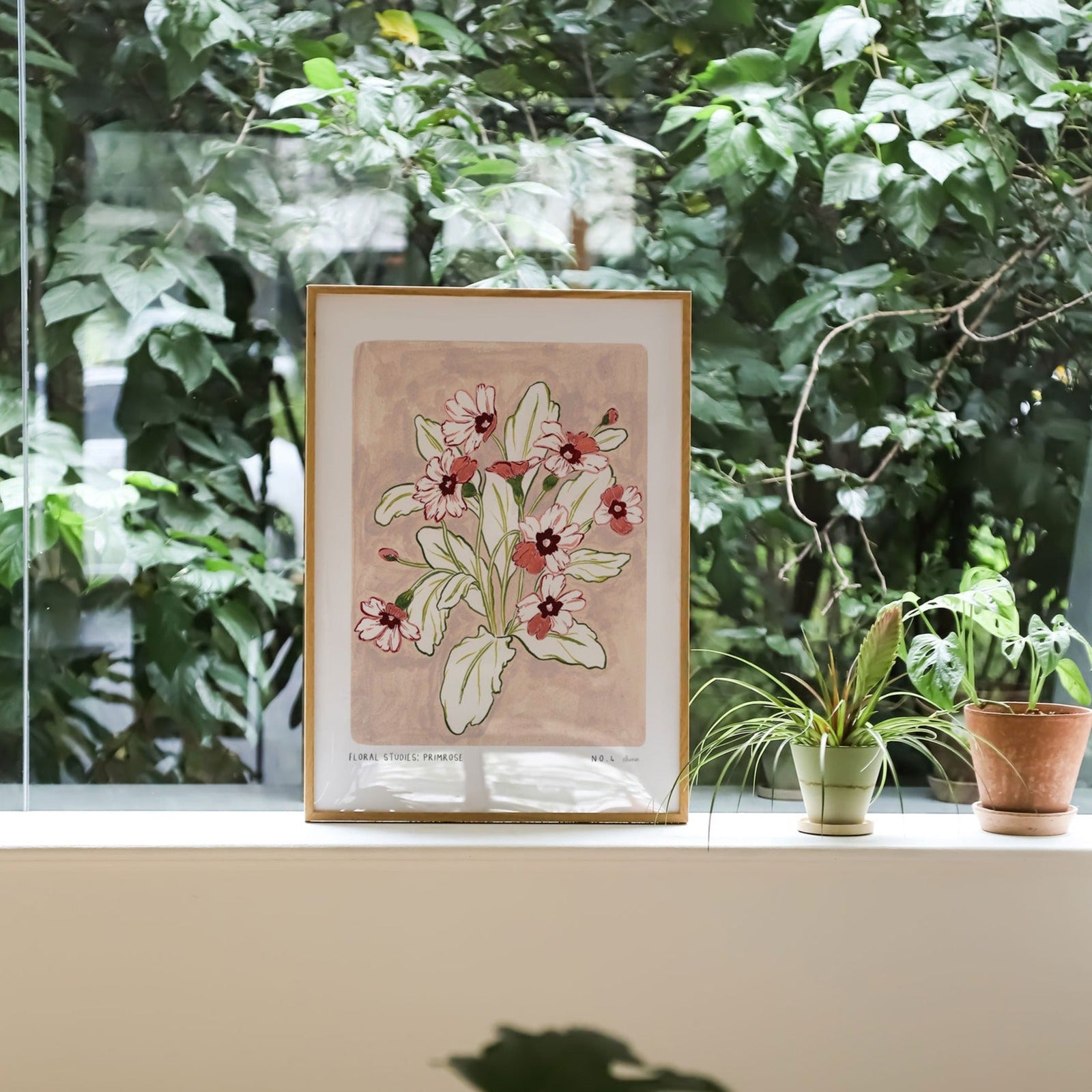 Primrose art print, a dusky light pink painted background with rosey pink and deep maroon flowers and olive green leaves. With small text reading floral studies:primrose NO.4 styled in a light wood frame against plants and greenery