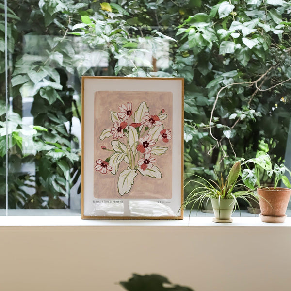 Primrose art print, a dusky light pink painted background with rosey pink and deep maroon flowers and olive green leaves. With small text reading floral studies:primrose NO.4 styled in a light wood frame against plants and greenery