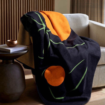 A dark navy throw blanket with abstract orange and green patterns is draped over a light beige armchair in a modern living room setting.