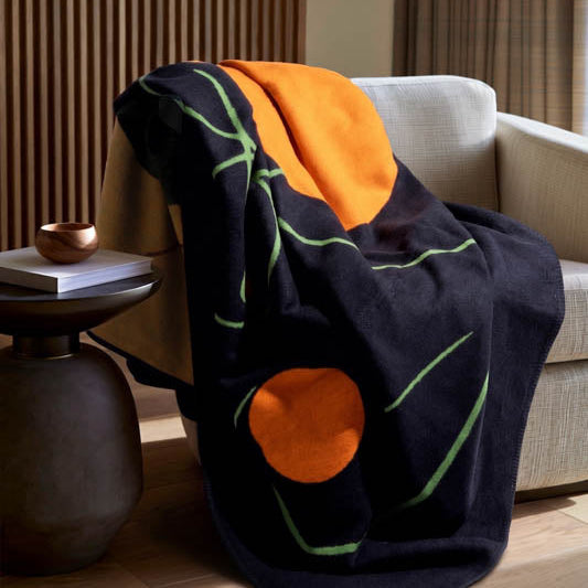 A dark navy throw blanket with abstract orange and green patterns is draped over a light beige armchair in a modern living room setting.