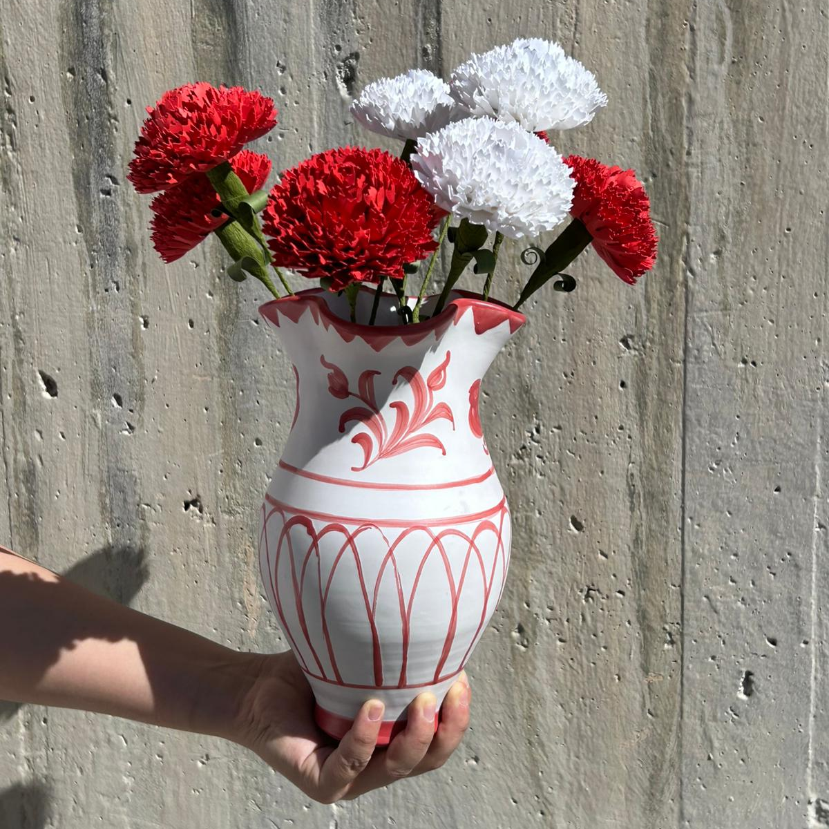 White vase with red floral patterns holding red and white flowers against a concrete wall. - lifestyle