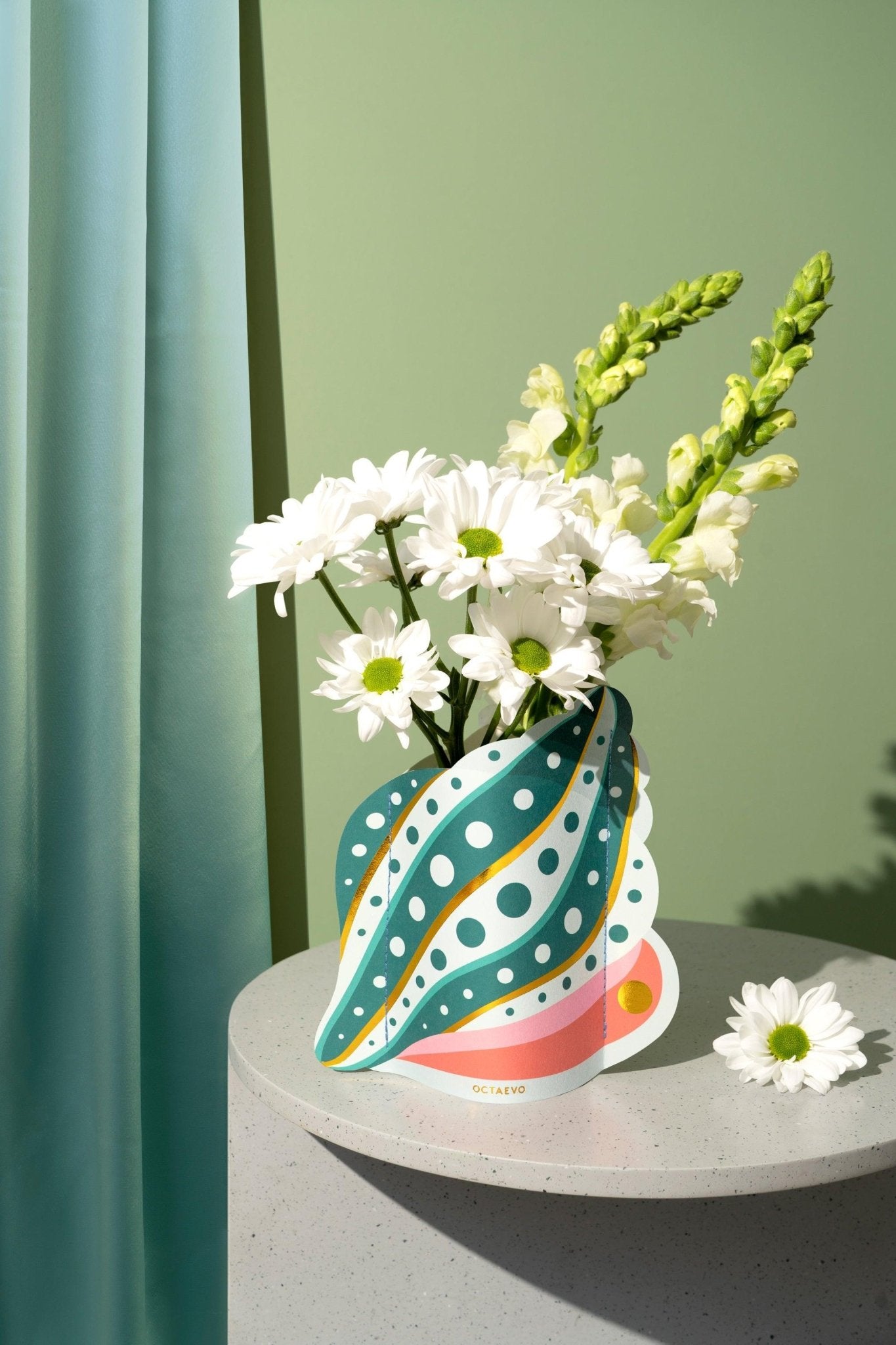 White daisies and snapdragons in the Octaevo Shell Mini Paper Vase, displayed on a gray table with a green background.