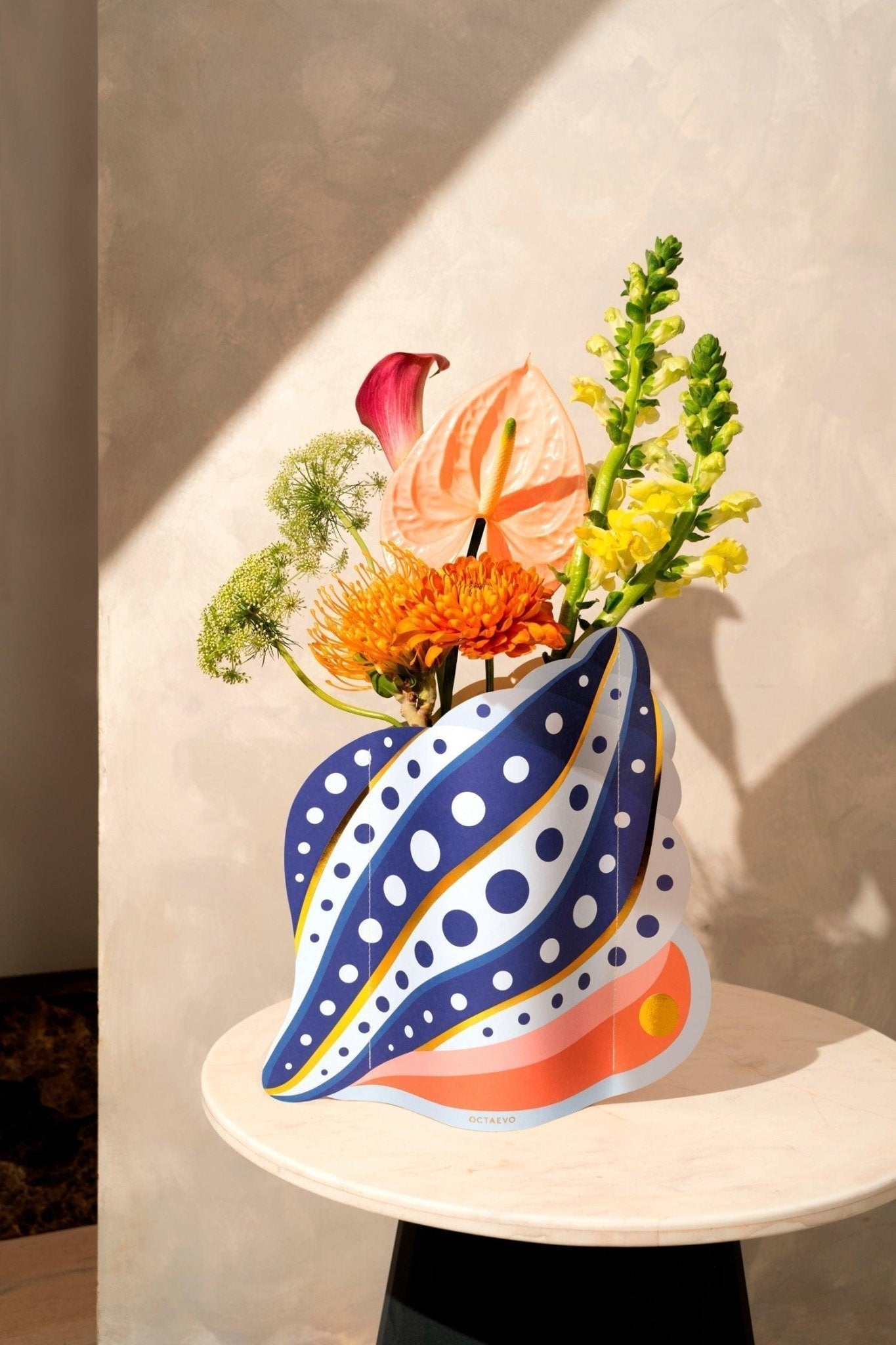 Vibrant flowers in Octaevos Shell Paper Vase create a Mediterranean centerpiece on a round table with a neutral backdrop.