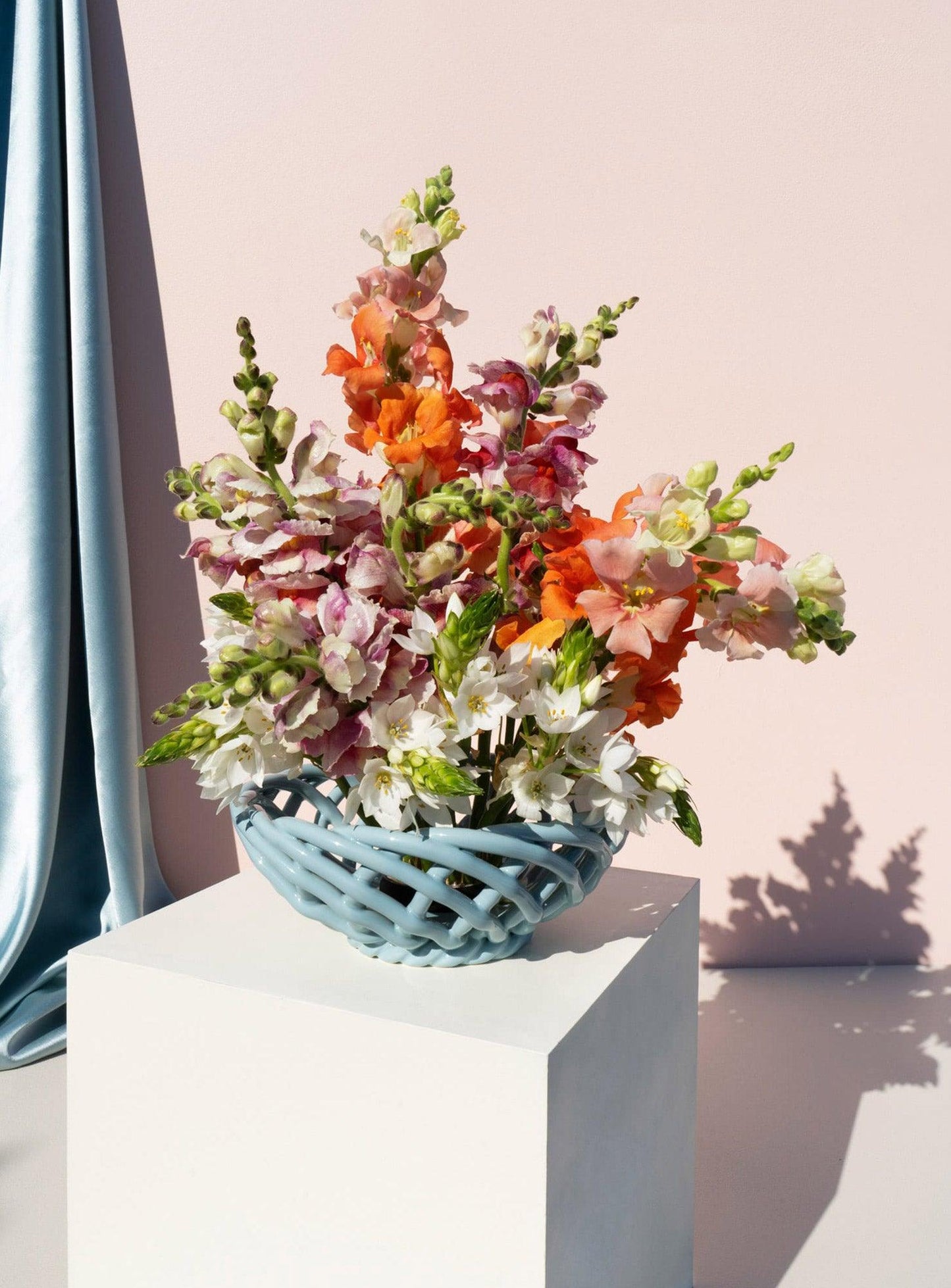 The light blue OCTAEVO Sicilia Ceramic Basket holds a vibrant bouquet on a white pedestal against a pink wall.