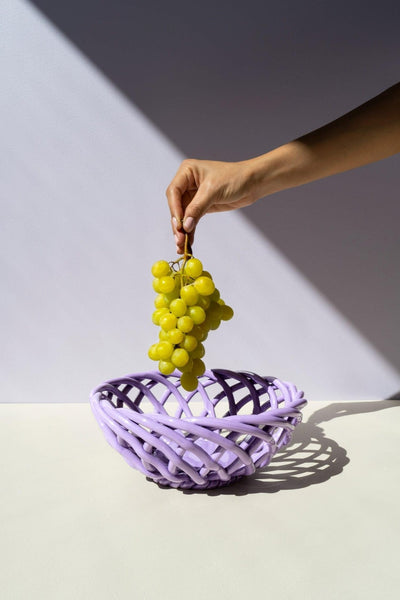 A hand holds green grapes above the purple OCTAEVO Sicilia Ceramic Basket on a white surface with sharp shadows.