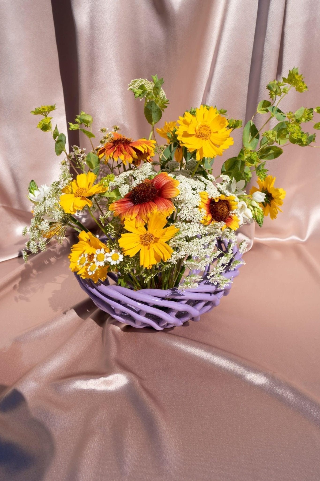 The purple Sicilia Ceramic Basket by OCTAEVO features yellow, orange, and white flowers with greenery on shiny pink fabric.