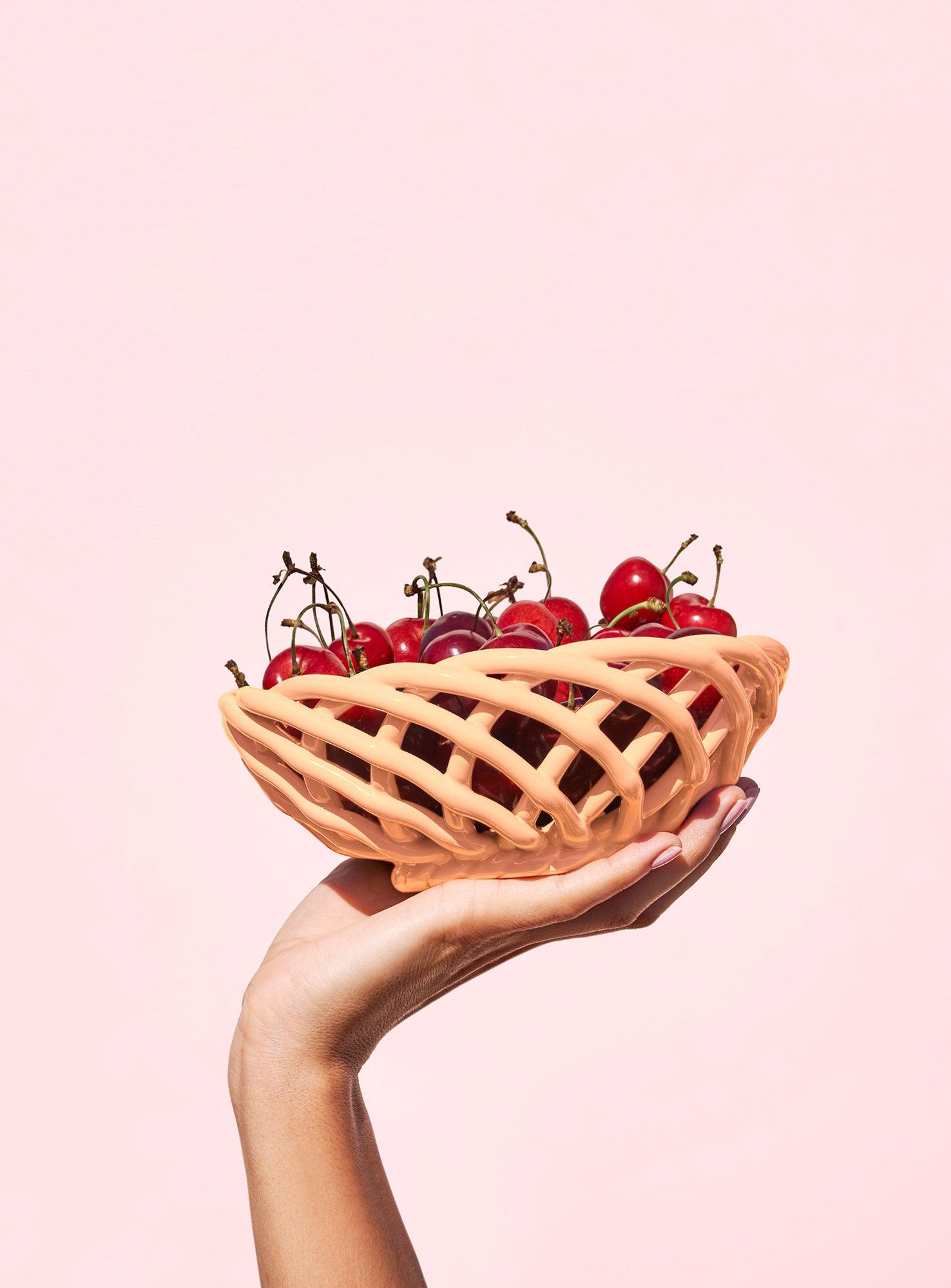 A hand holds an orange OCTAEVO Sicilia Ceramic Basket filled with fresh cherries against a light pink background.