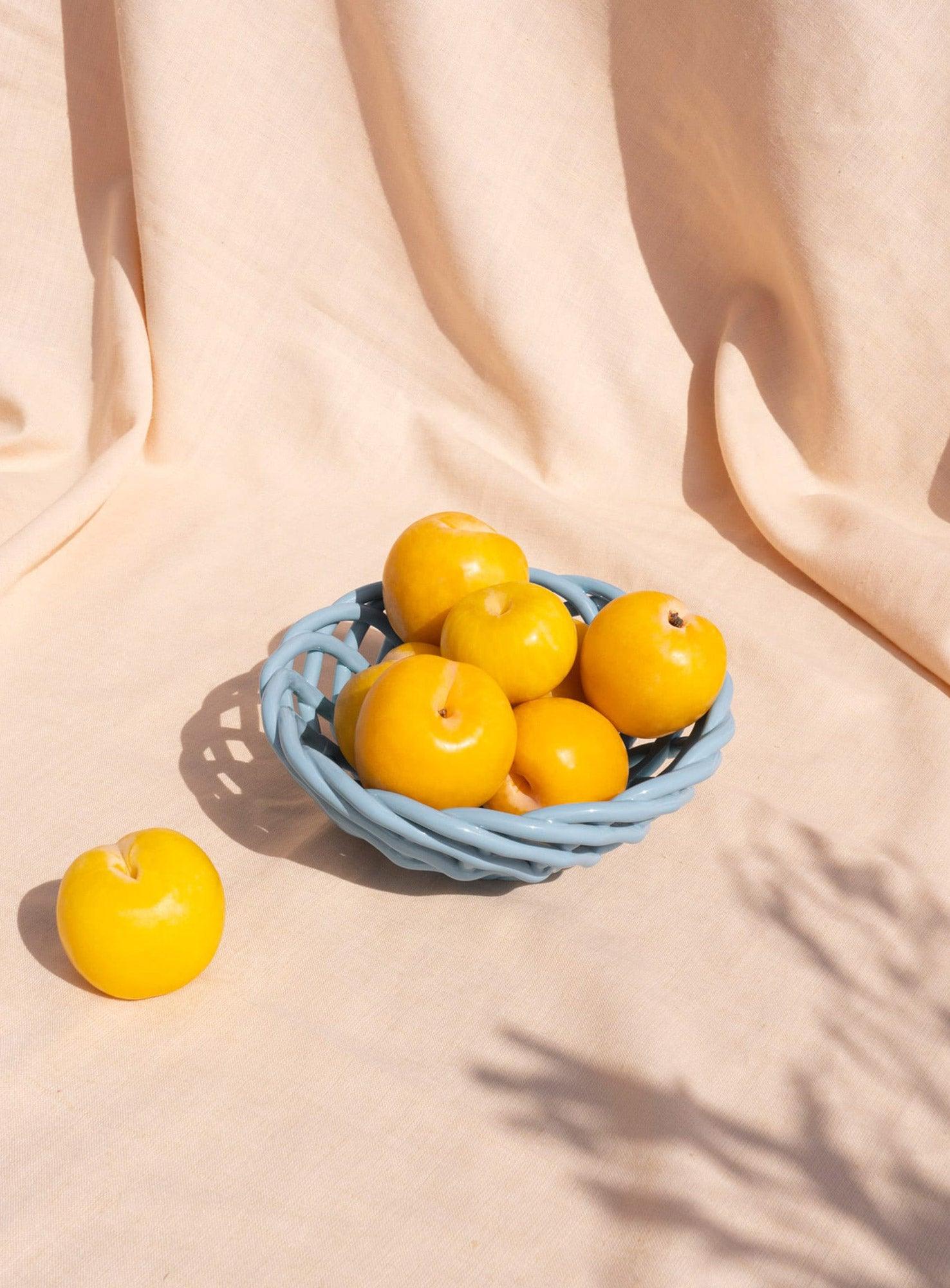 The light blue OCTAEVO Sicilia Ceramic Basket holds yellow plums on a beige cloth. 