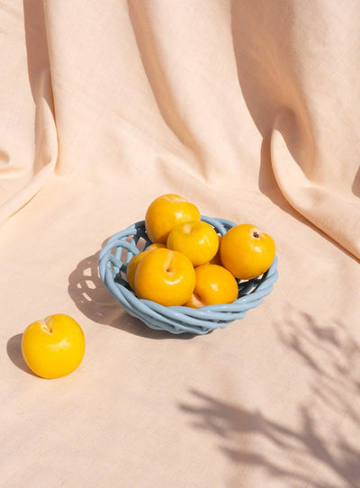 The light blue OCTAEVO Sicilia Ceramic Basket holds yellow plums on a beige cloth. 