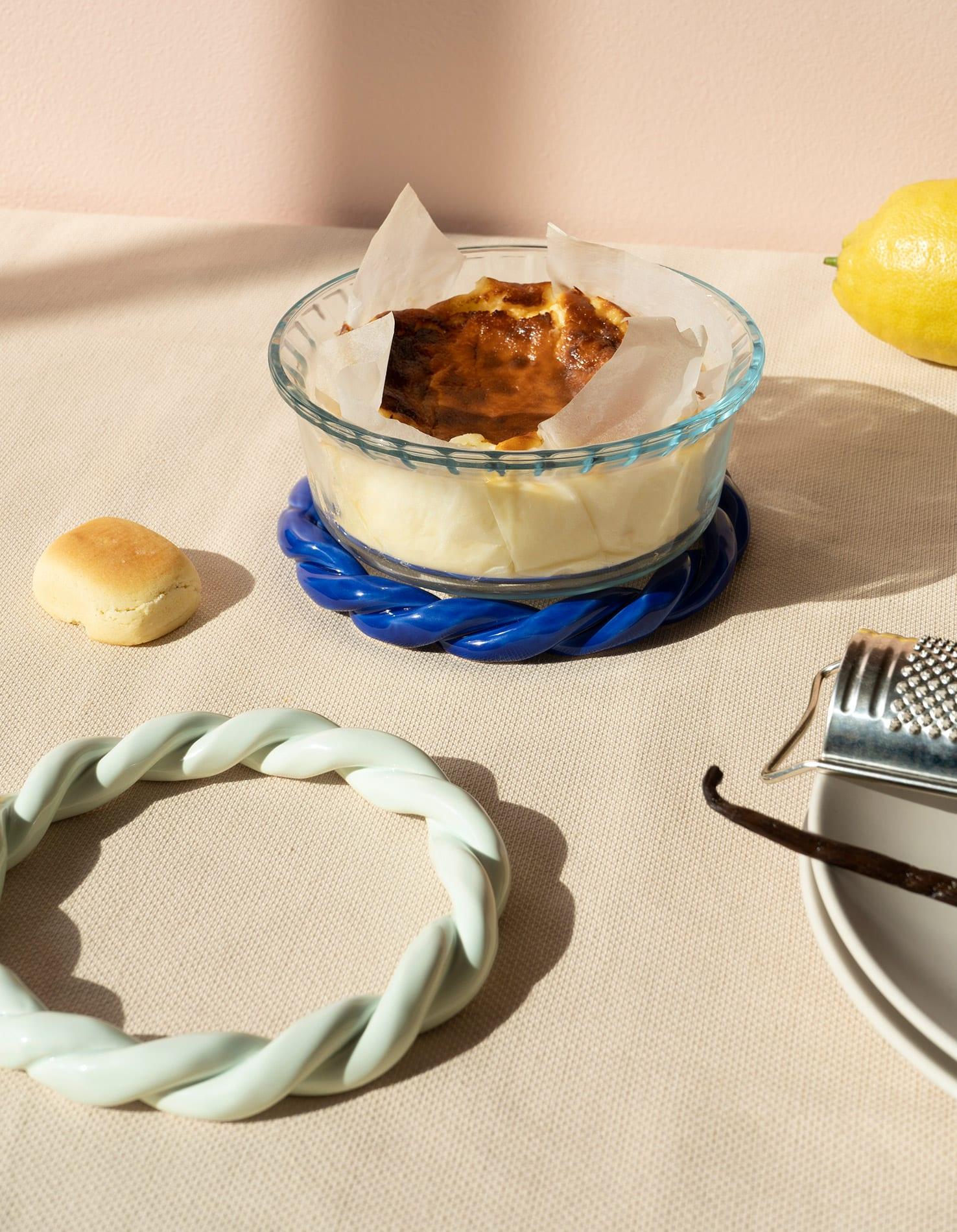 A baked dish in a glass bowl rests on an blue OCTAEVO Sicilia Ceramic Trivet, with lemon, bread roll, grater, and vanilla bean.