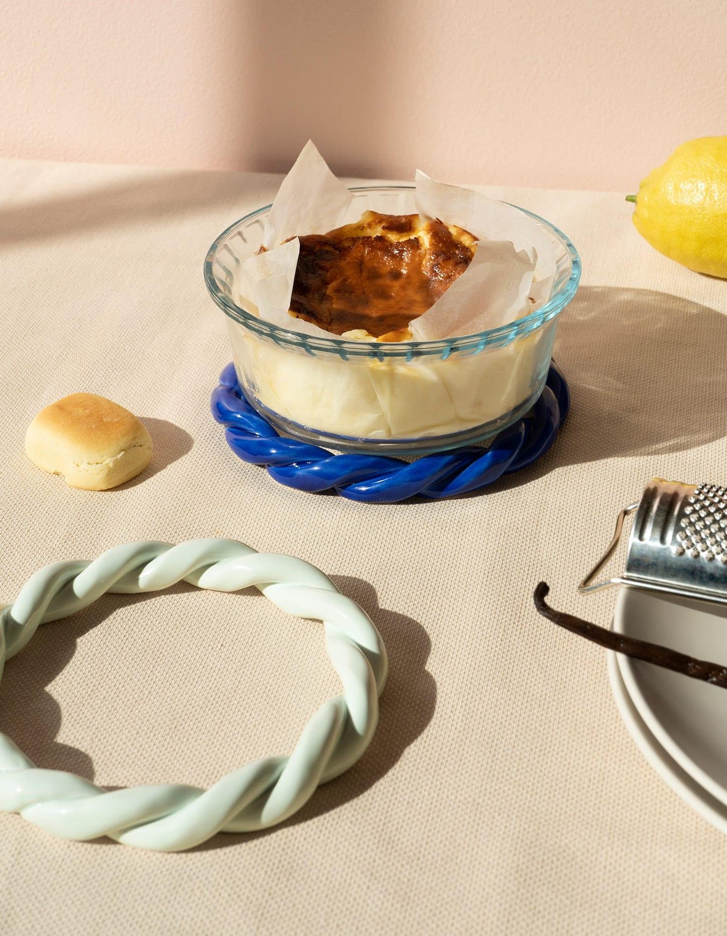 A baked custard dessert in a glass dish on a light mint trivet, with a lemon, a roll, and a vanilla bean nearby.